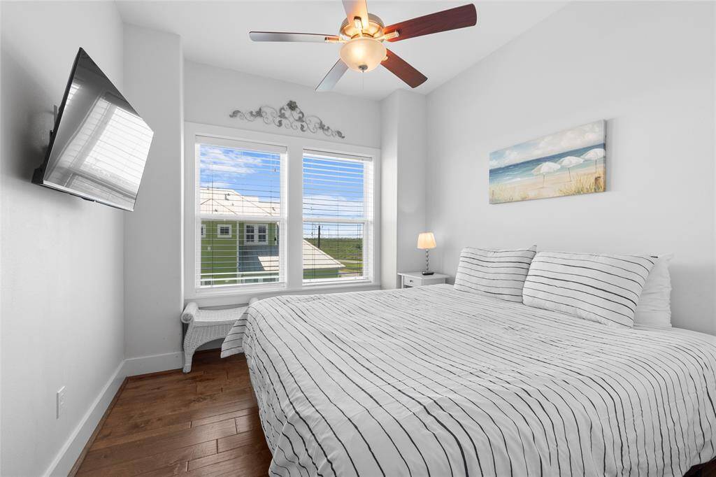 Bright bedroom with a cozy bed, featuring striped bedding and a ceiling fan. Large windows provide natural light and a view. The room includes a wall-mounted TV and beach-themed decor, creating a relaxing atmosphere.