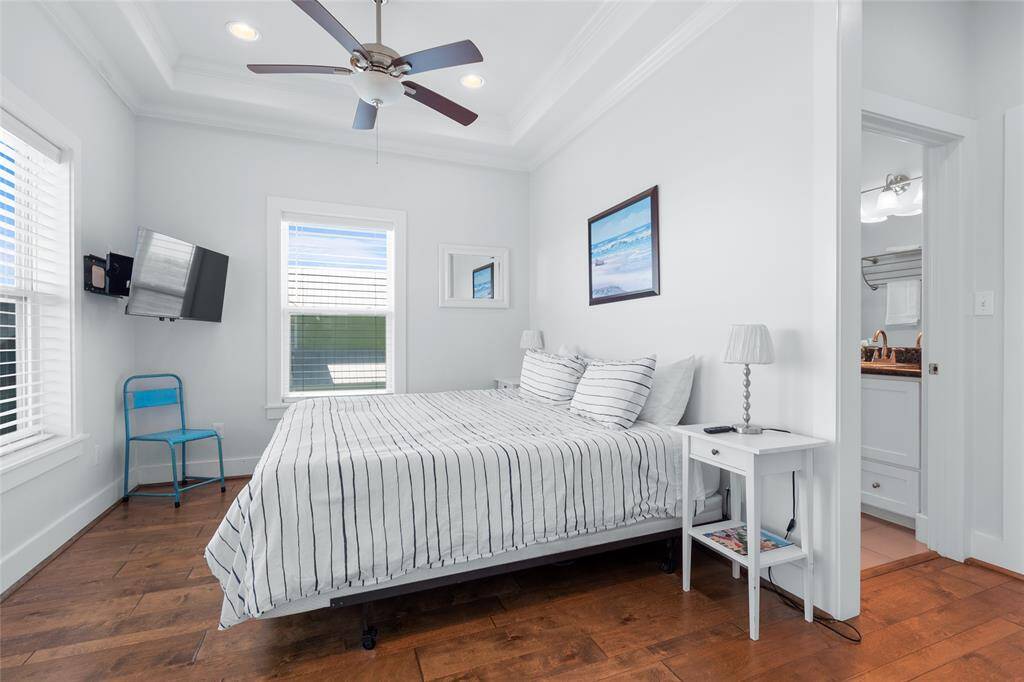 Bright and inviting bedroom with hardwood floors, featuring a ceiling fan, wall-mounted TV, and ample natural light. The room is furnished with a cozy bed and nightstands, and includes access to an ensuite bathroom.