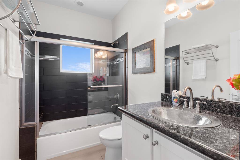 This bathroom features a modern design with black tile accents, a glass-enclosed shower-tub combo, and a granite countertop with a stylish sink. Bright lighting and a large mirror enhance the space, while a towel rack and decorative touches add functionality and charm.