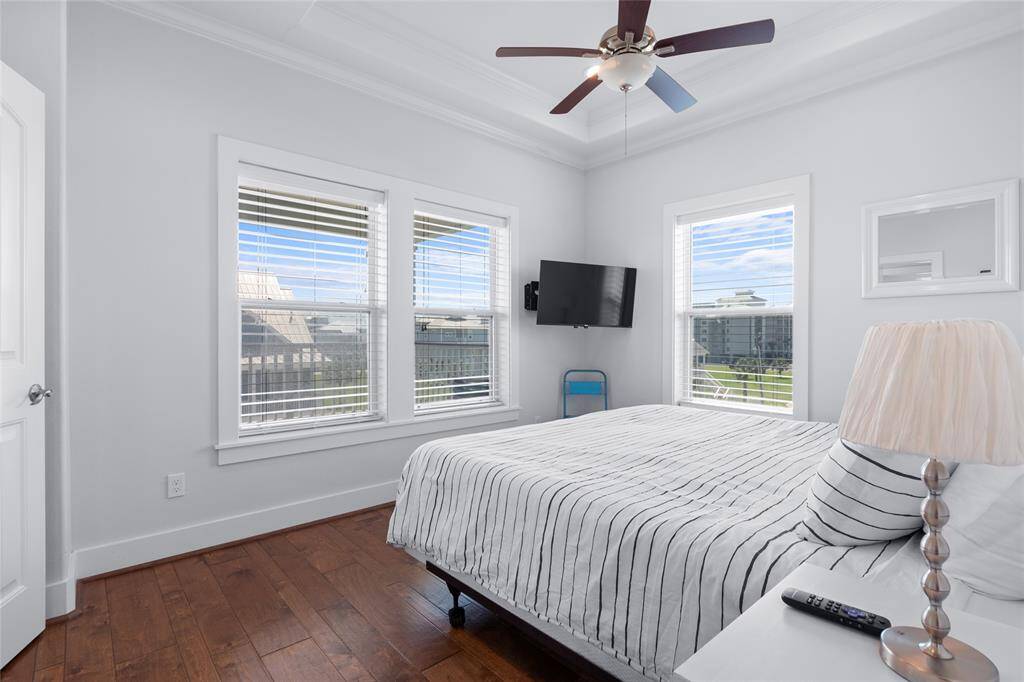 Bright bedroom with hardwood floors and a ceiling fan. It features large windows for natural light, a wall-mounted TV, and a neatly made bed with striped bedding.