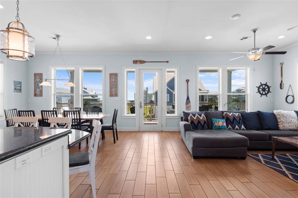 This photo showcases a bright, open-concept living and dining area with large windows and glass doors offering plenty of natural light. The space features a modern kitchen with a granite countertop, a spacious dining area with a large table, and a cozy living room with a sectional sofa. Nautical decor accents add charm and character.
