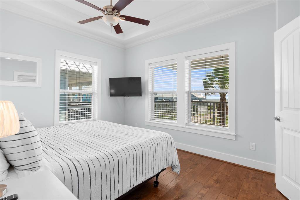 This bright bedroom features hardwood floors, a ceiling fan, and a wall-mounted TV. Large windows provide natural light and a view, creating an inviting and cozy atmosphere.