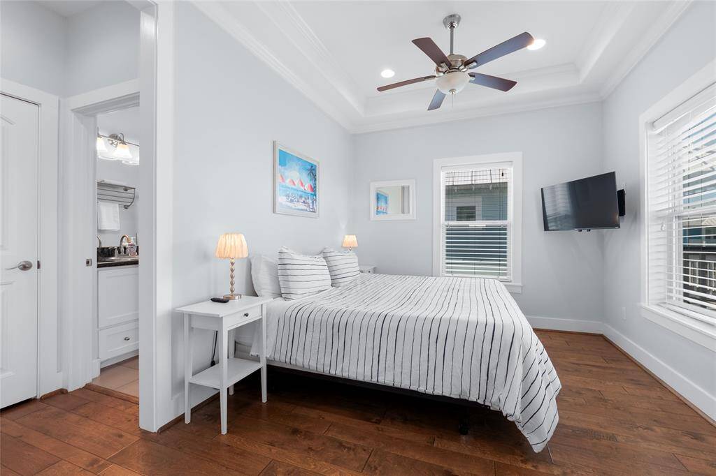 Bright, modern bedroom with hardwood floors, featuring a ceiling fan, wall-mounted TV, and an en-suite bathroom. The room has large windows and a cozy, inviting atmosphere.