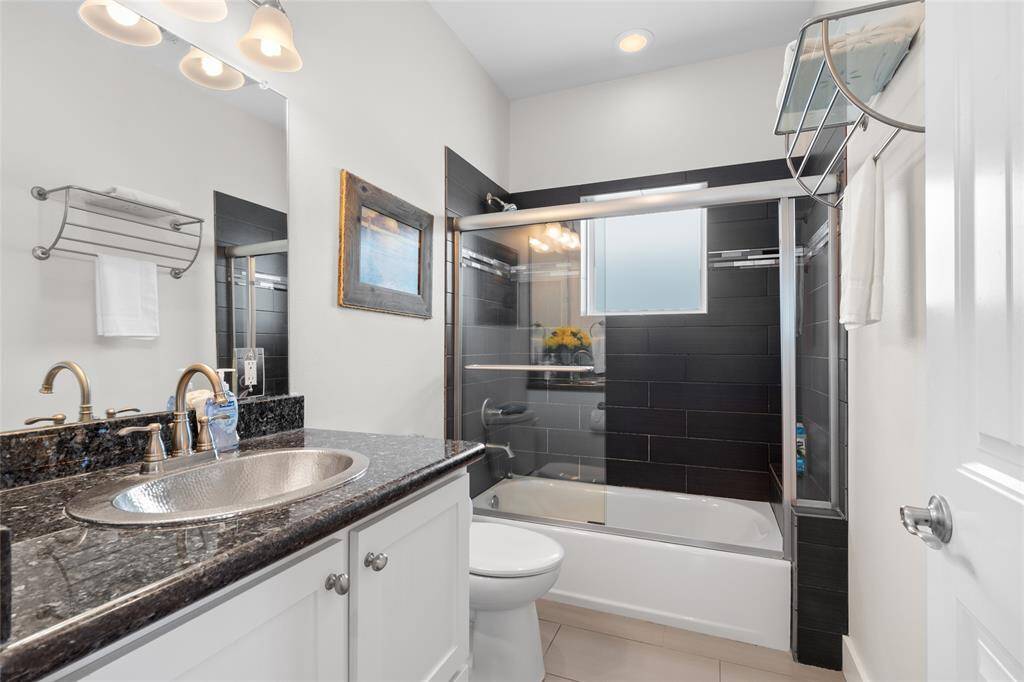 This bathroom features a sleek design with a granite countertop and a modern vessel sink. It includes a shower-tub combo with stylish black tile and a glass sliding door. The space is well-lit with overhead lighting and has a contemporary feel.
