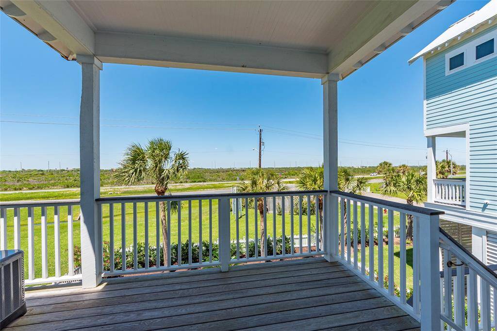 This photo showcases a charming covered porch with wooden flooring, offering a picturesque view of a lush green lawn and palm trees. The porch is part of a coastal-style home, perfect for enjoying serene outdoor moments.