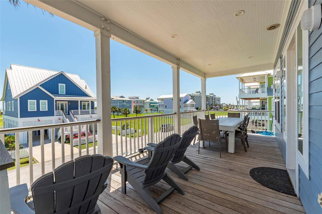 This photo showcases a spacious, covered porch with comfortable seating and a dining table, offering views of neighboring coastal homes in a bright, well-maintained neighborhood. Ideal for relaxing and entertaining.