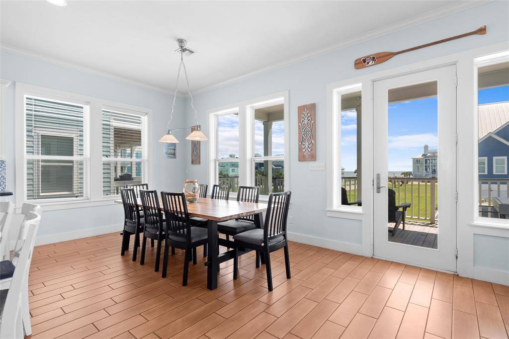 This bright dining area features a spacious table set against a backdrop of large windows, offering abundant natural light and a view of the outdoors. The room has a coastal theme with light blue walls, wood-style flooring, and access to a balcony, making it perfect for entertaining or family meals.