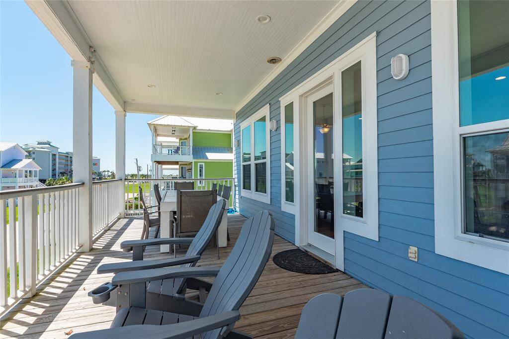 This photo shows a charming porch with a coastal vibe, featuring blue siding and white trim. It offers comfortable seating and a dining area, overlooking a neighborhood with colorful houses. Perfect for relaxing and enjoying the view.