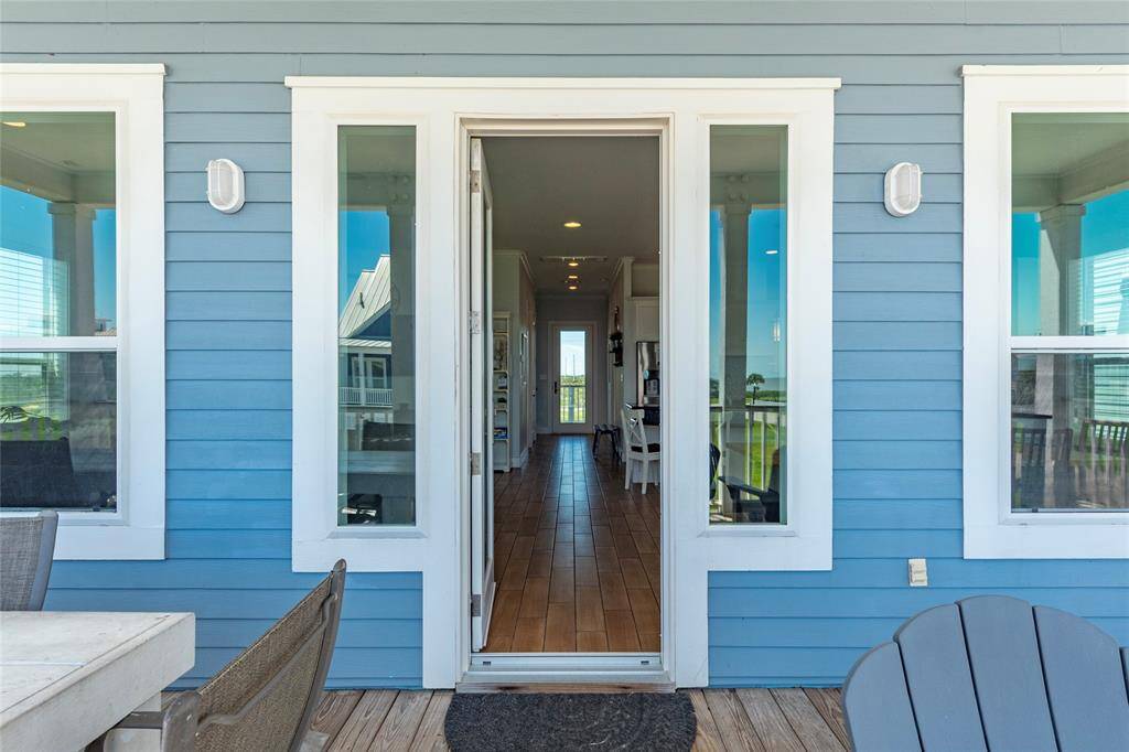 This photo showcases a charming entryway with a light blue exterior and white trim, leading to an inviting interior through a glass door. Flanked by large windows, it opens to a cozy deck area with outdoor seating, offering a glimpse of a spacious and bright interior.