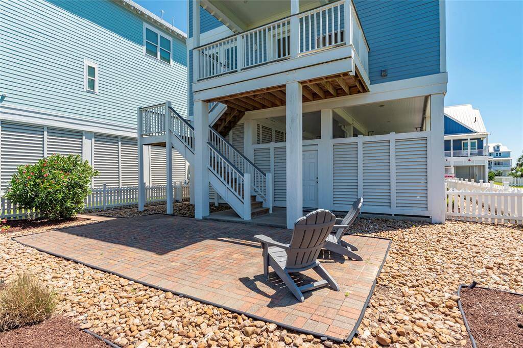 Charming coastal-style home with a raised patio area featuring Adirondack chairs, accessible by stairs. The backyard is landscaped with stone paths and a small garden, perfect for outdoor relaxation.