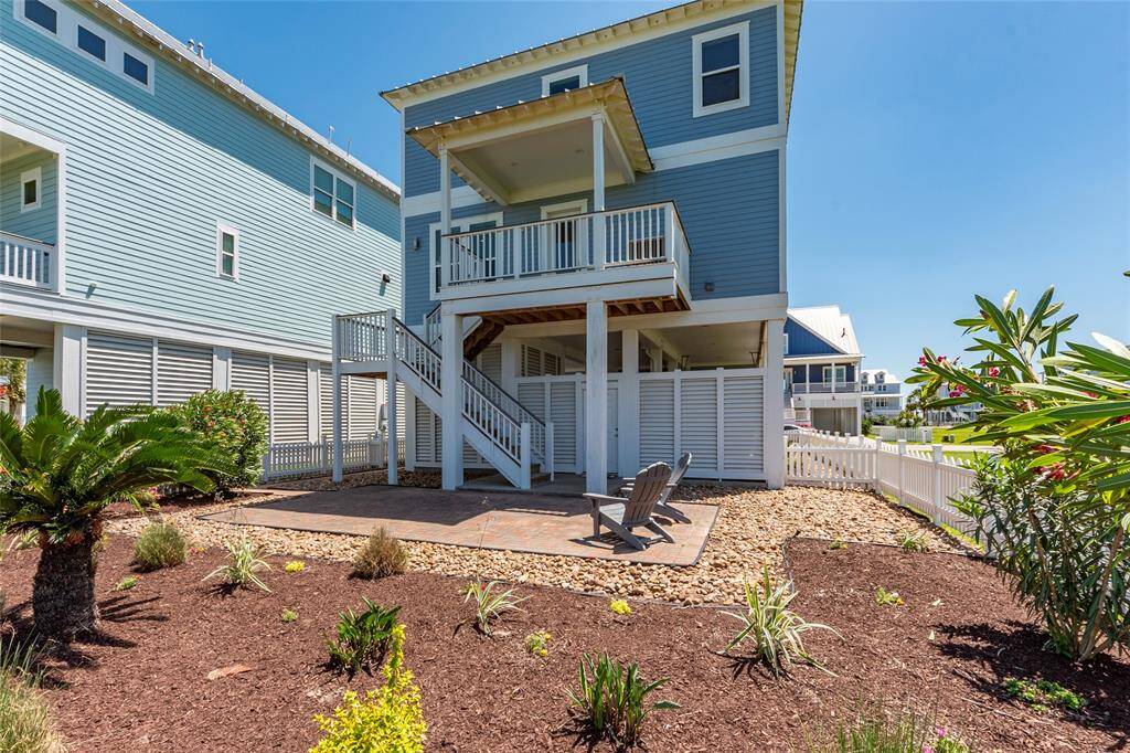 This photo shows a modern, two-story home with a blue exterior and white trim, featuring a covered balcony and an exterior staircase. The backyard has a patio area with seating, surrounded by landscaped gardens and a white picket fence.