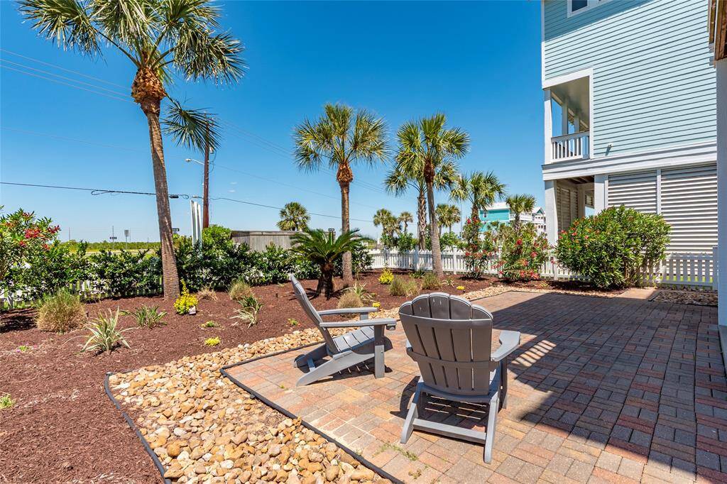 This photo showcases a well-maintained backyard with a paved patio, featuring two Adirondack chairs. The area is surrounded by lush landscaping, including palm trees and various shrubs, creating a relaxed, tropical atmosphere.
