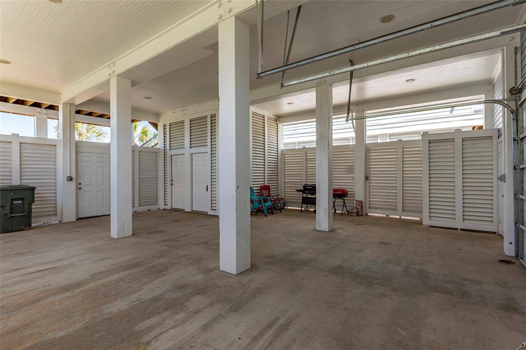 This photo shows a spacious covered garage area with concrete flooring and white louvered walls. It includes a few storage areas and a utility door, providing ample space for parking and additional storage.