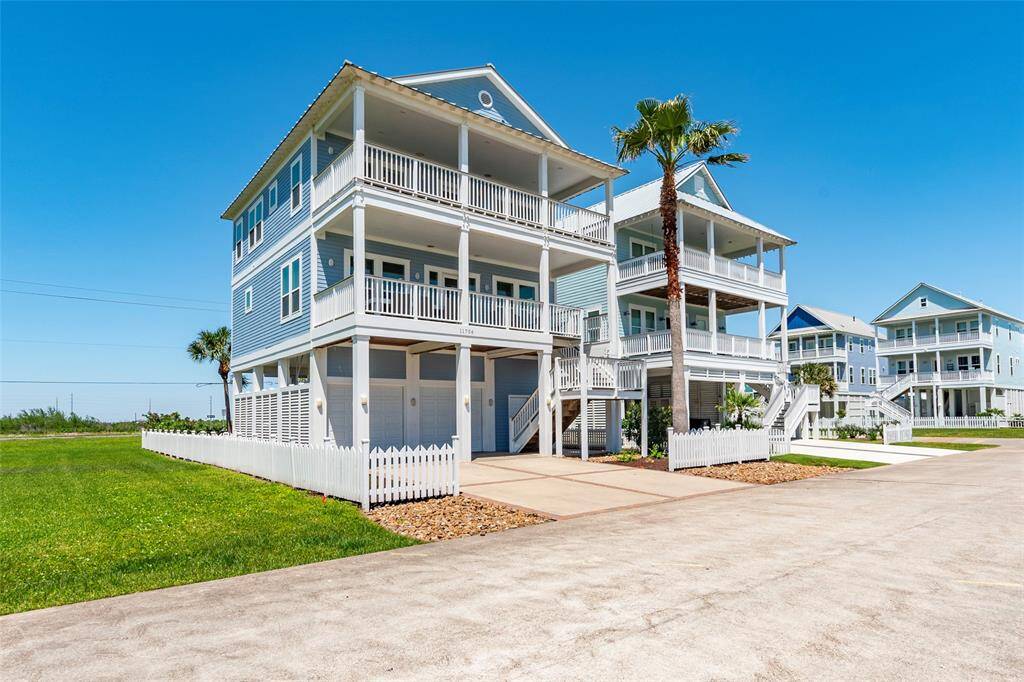 This is a spacious, multi-story coastal home with a classic blue exterior and white trim. It features multiple balconies, a fenced yard, and palm trees, offering a charming seaside vibe. The neighborhood appears well-maintained and inviting.