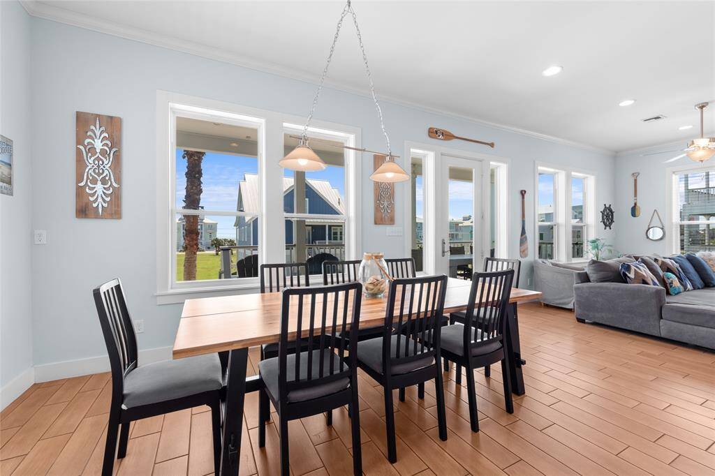 This bright, open-concept living and dining area features large windows offering plenty of natural light and views of the outdoors. It includes a spacious dining table, modern light fixtures, and comfortable seating, set against a backdrop of light-colored walls and wood-style flooring.