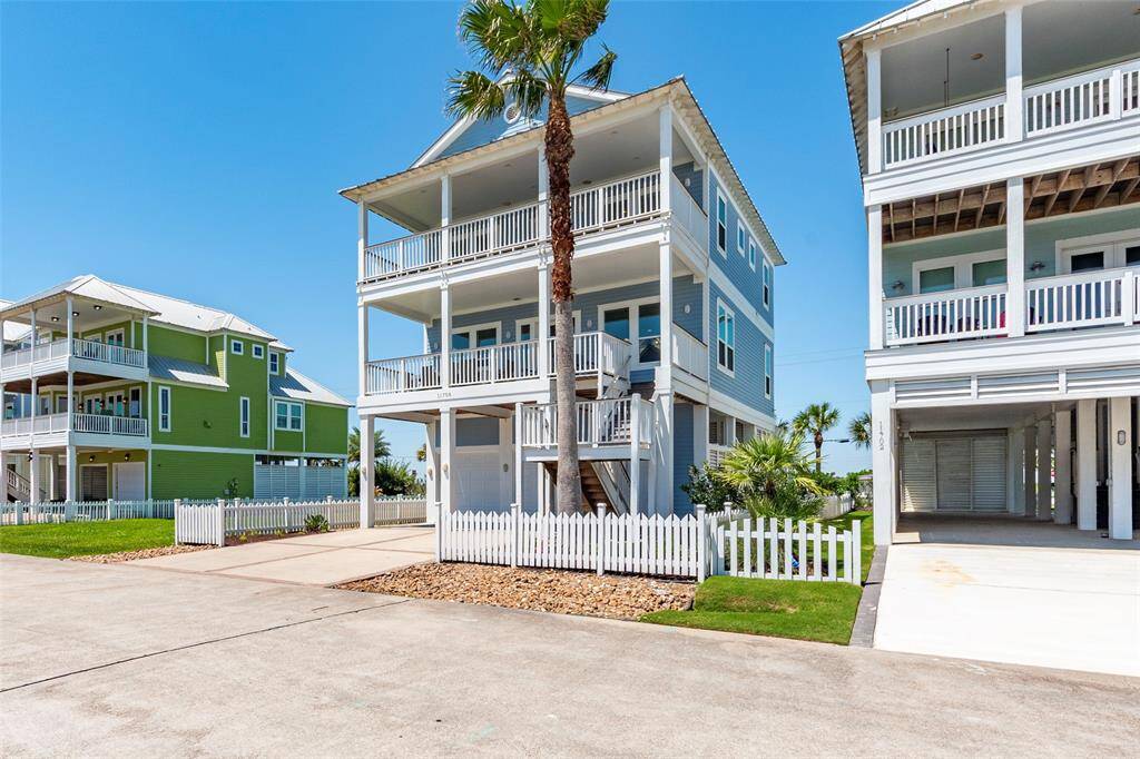 This image features a modern, three-story coastal home with a light blue exterior and spacious balconies. It is flanked by similar houses, each with white railings and a small fenced yard. The property includes a driveway and a palm tree, contributing to a beachside atmosphere.