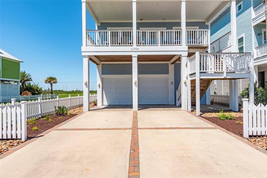 This photo shows a coastal-style home with an elevated structure, featuring a spacious two-car garage and a welcoming front porch. The home is surrounded by a white picket fence and neatly landscaped gardens, offering a charming and well-maintained exterior.