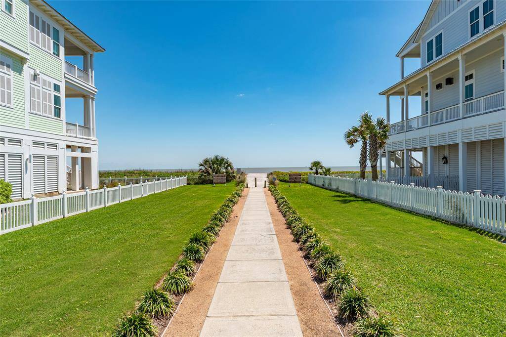 This photo showcases a beautiful coastal property with a paved walkway leading to the beach. Flanked by two charming homes with wraparound porches, the scene features well-maintained lawns and white picket fences, offering a serene, inviting atmosphere.