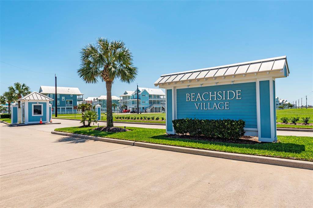 This photo showcases the entrance to Beachside Village, featuring a welcoming sign, a small security booth, and well-maintained landscaping with palm trees, set against a backdrop of coastal-style homes under a clear blue sky.