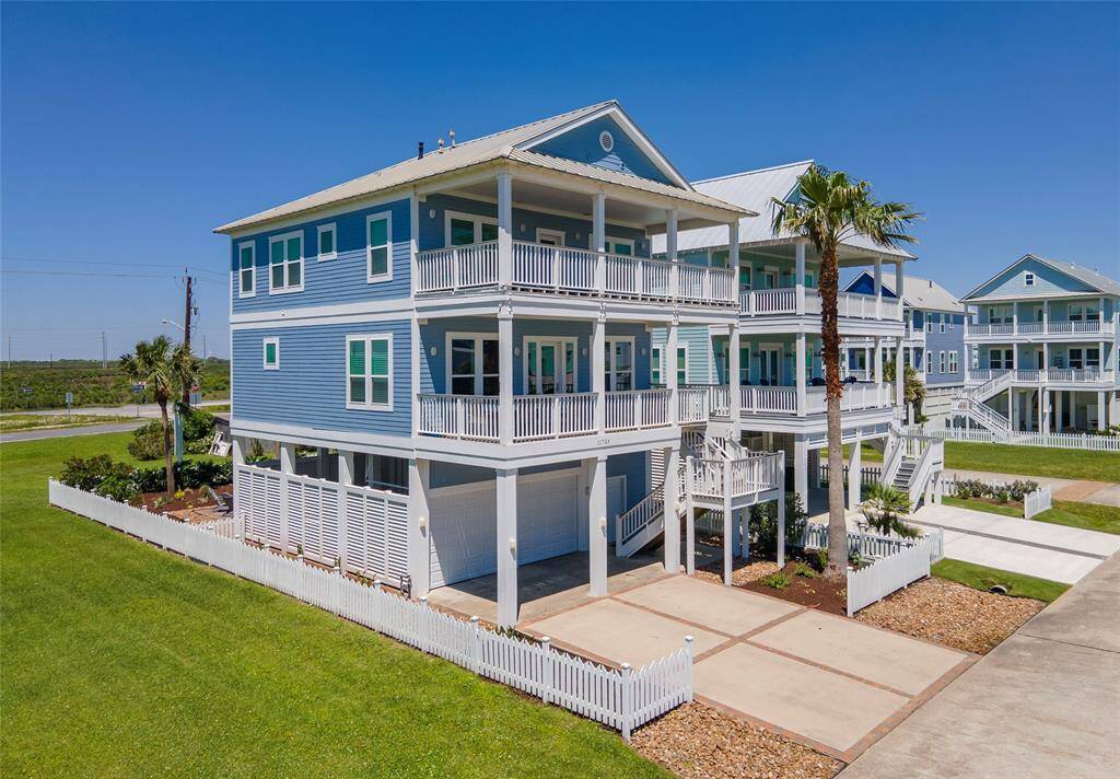 This is a charming coastal home with a bright blue exterior and multiple balconies, offering beautiful views. It features a white picket fence, a spacious driveway, and a well-maintained lawn, creating a welcoming atmosphere.