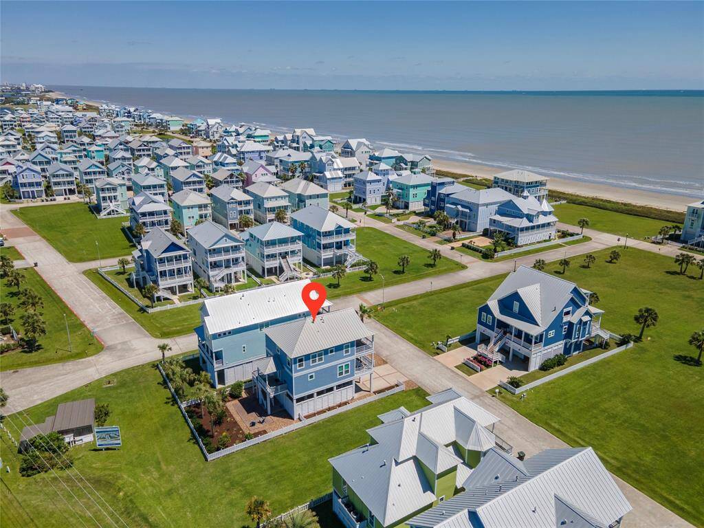 This aerial photo showcases a coastal neighborhood with colorful, elevated homes near the beach. The highlighted house features a modern design with ample outdoor space, offering proximity to the ocean and scenic views. Ideal for those seeking a beachside lifestyle in a vibrant community.