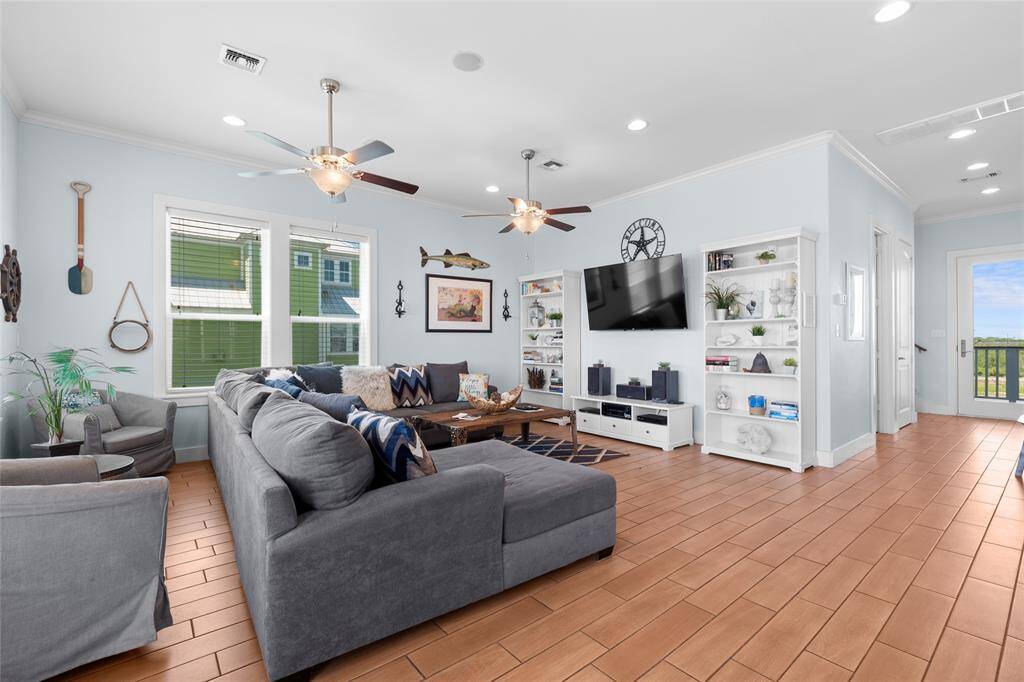 Bright and inviting living room with light blue walls and wood-look tile flooring. Features a cozy sectional sofa, ceiling fans, large windows, and built-in shelving. Wall-mounted TV and decorative accents add a modern touch. Adjacent door leads to outdoor space.