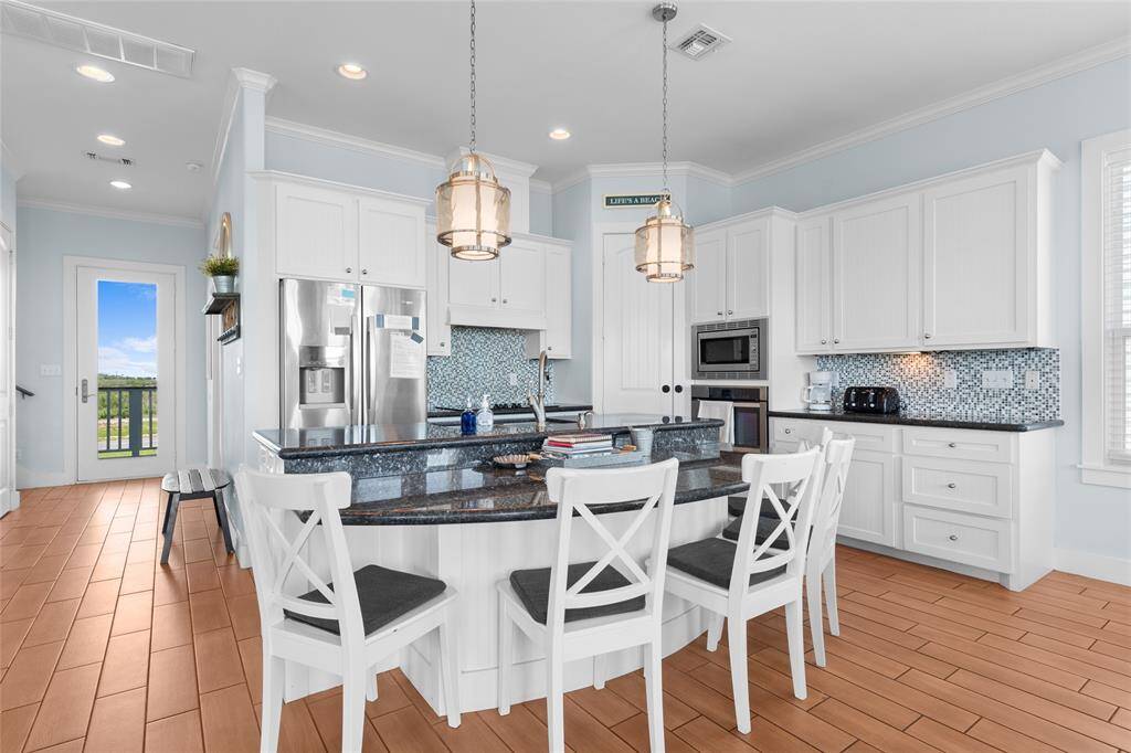 This kitchen features modern white cabinetry, a stylish mosaic tile backsplash, and stainless steel appliances. It includes a large island with seating for four, pendant lighting, and wood-style flooring. Natural light streams through windows and a glass door leading to an outdoor space.