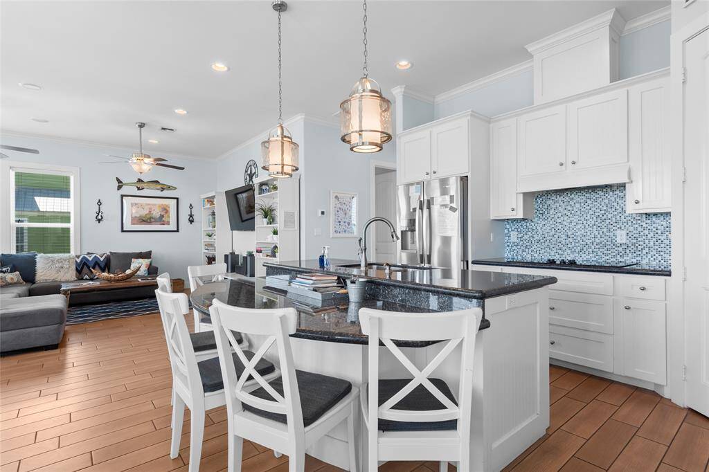 This open-concept living space features a modern kitchen with white cabinetry, a stylish blue mosaic backsplash, and a sleek black countertop island with seating. The adjoining living area is cozy, with a sectional sofa and tasteful decor, creating a welcoming atmosphere. Natural light enhances the bright and airy feel.