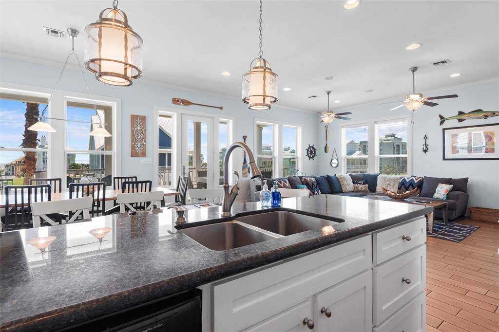 The photo showcases a bright, open-concept living area with coastal decor. It features a spacious kitchen island with a sink, modern pendant lighting, and a large dining area. The room has ample natural light from multiple windows and a cozy seating area, perfect for entertaining.