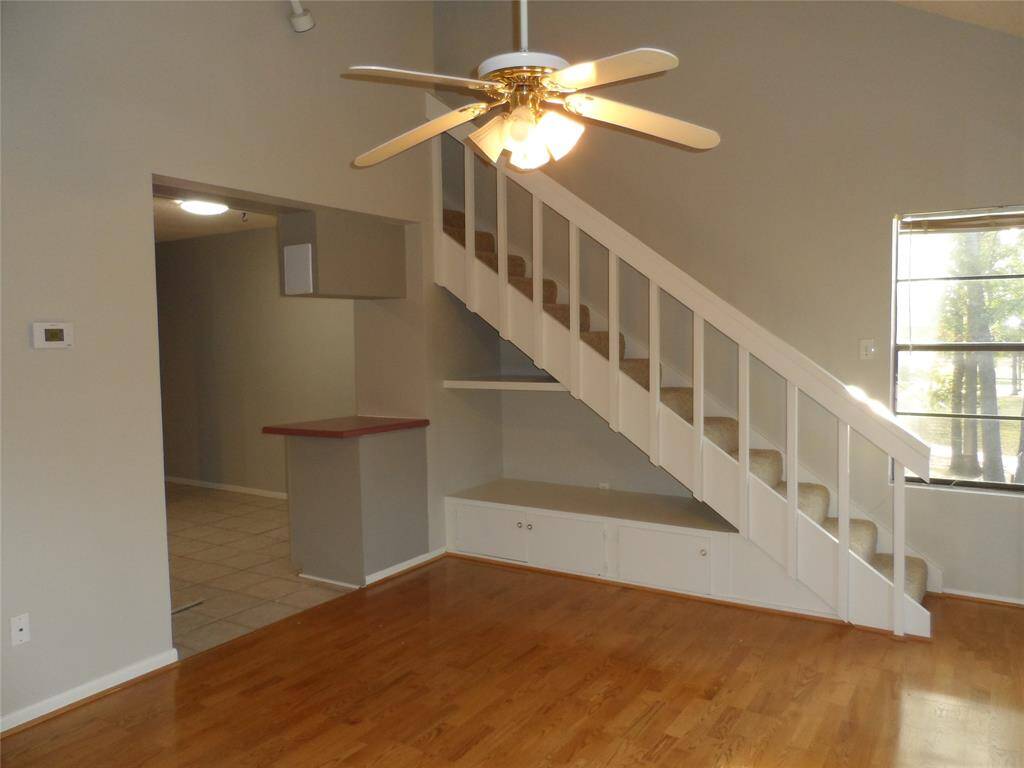 Cozy living area with hardwood like floors, a ceiling fan, and an open staircase with storage underneath. The space connects to a tiled area leading to the kitchen. Natural light comes through the window, enhancing the room's warmth.