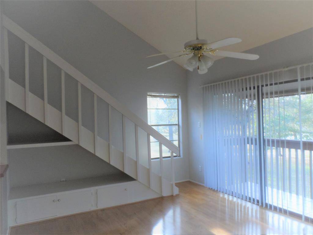 Bright living space with high ceilings features a ceiling fan and large windows with vertical blinds. The room includes a staircase with built-in storage underneath, and light wood like flooring.