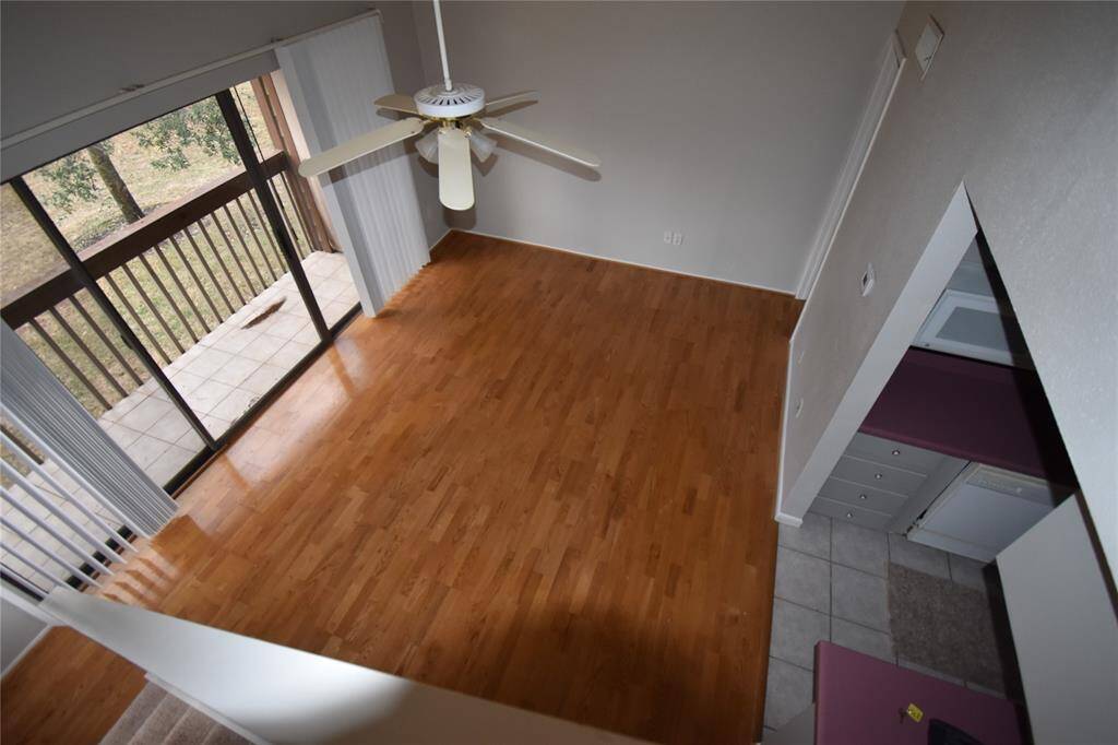 Spacious living area with wood like floors, a ceiling fan, and sliding glass doors leading to a balcony with a view of greenery. Adjacent is a tiled kitchen area with visible cabinetry.
