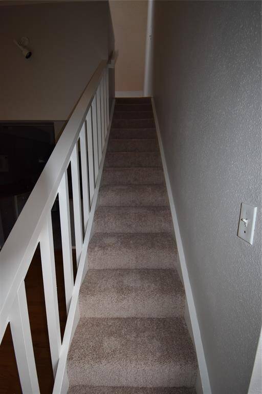 This photo shows a carpeted staircase with a white railing and neutral wall colors, leading to an upper floor.