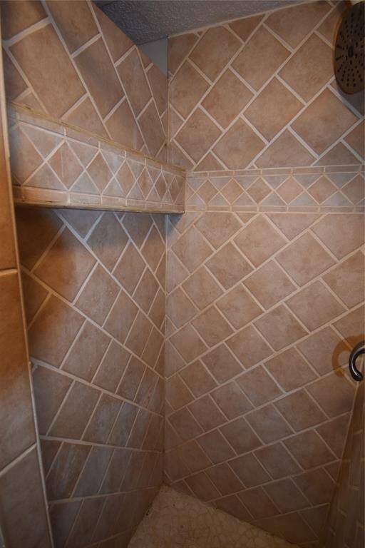 This photo shows a shower with beige diagonal tile walls and a built-in shelf. The showerhead is mounted on the right, and the floor has small pebble-like tiles, adding texture and contrast.