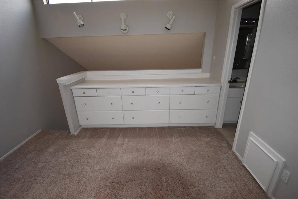 This room features a built-in white dresser with ample storage and overhead lighting. It has neutral carpeting and direct access to an adjoining bathroom, making it a practical and functional space.