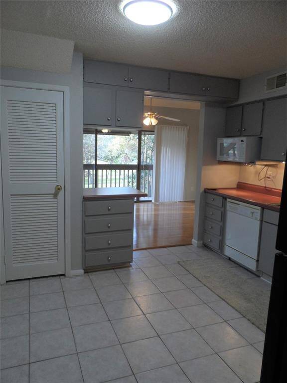 The kitchen with gray cabinets, tiled flooring, and ample natural light. It opens into a dining area with hardwood like floors and large windows overlooking a balcony.