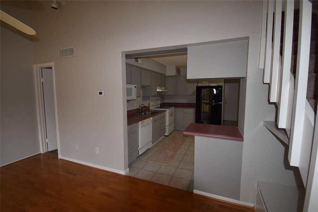 This photo shows an open-plan kitchen with a view from the living area. The kitchen features light cabinetry, and essential appliances like a refrigerator and microwave. It opens to a space with hardwood like flooring and a staircase, creating a cozy, functional layout.