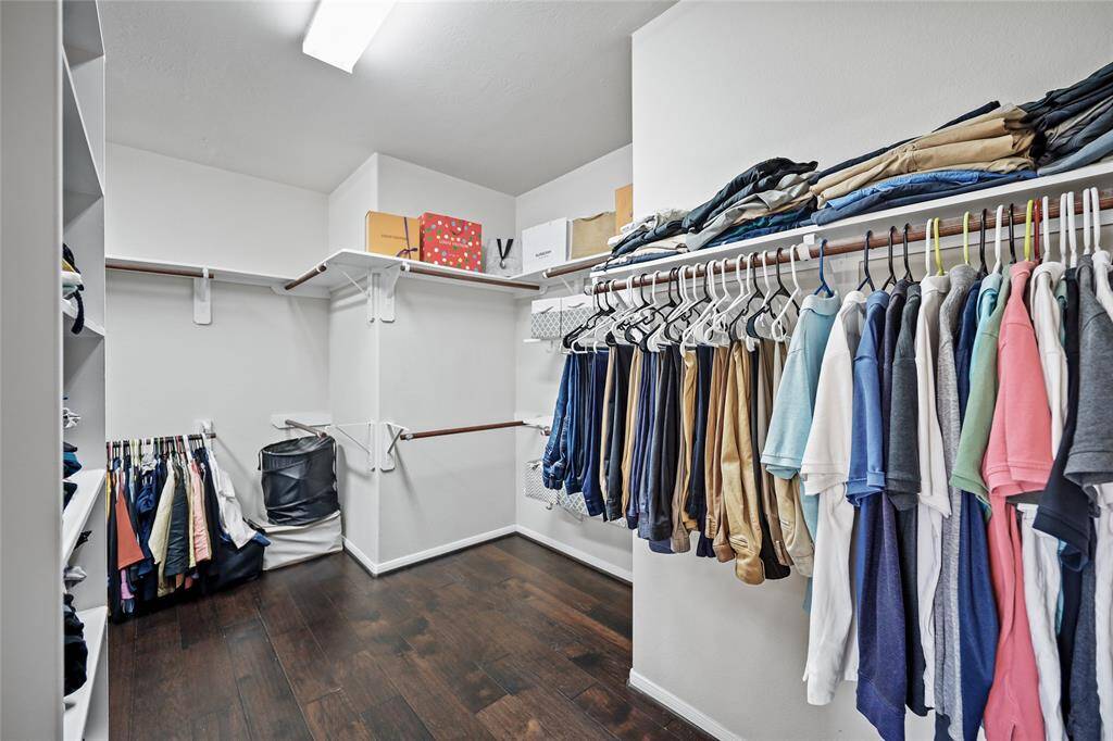 This spacious walk-in closet features dark wood flooring, ample hanging space with organized clothing racks, and overhead shelves for additional storage. It's well-lit and offers a neat, functional layout.
