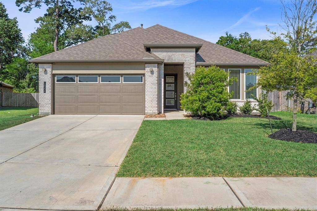 A beautifully maintained front elevation showcases a modern blend of light-toned brick and clean architectural lines. The covered entryway features a contemporary glass-panel front door with a recessed arch for added dimension. A well-manicured lawn, neatly mulched flower beds, and a double-wide driveway lead to the two-car garage with sleek horizontal paneling and clerestory-style windows.