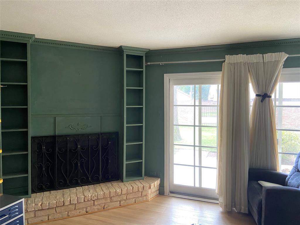 A cozy living area featuring a green built-in shelving unit with a decorative fireplace and wrought iron screen. The room has natural light from a large sliding glass door with sheer curtains, leading to an outdoor space. The brick hearth adds a classic touch.