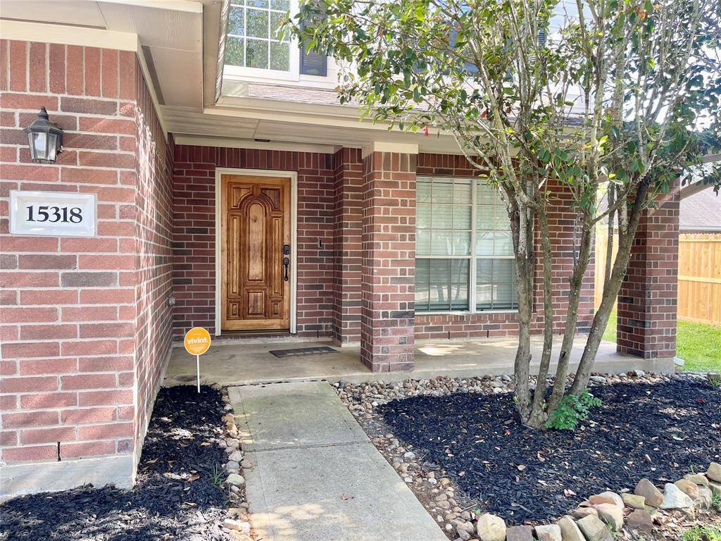 Charming brick home with a welcoming front porch and elegant wooden door. The entrance is framed by a neat garden area with mature trees and fresh landscaping. A security sign adds a sense of safety.
