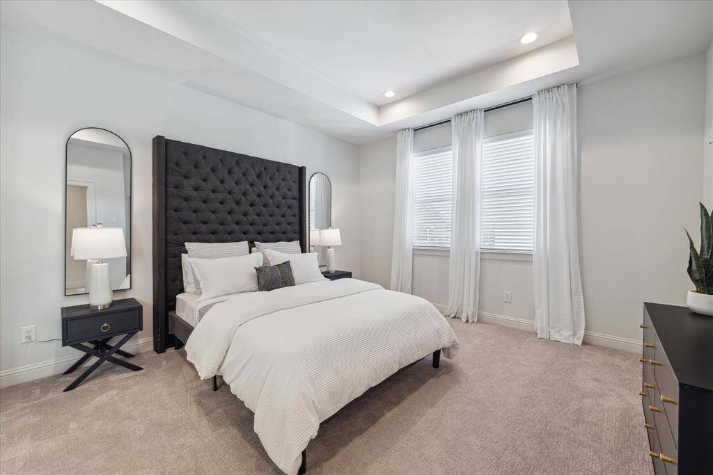 This spacious primary bedroom offers a serene retreat with soft carpeting and large windows that flood the room with natural light. The recessed ceiling and neutral color palette create a soothing and airy ambiance, perfect for relaxation.