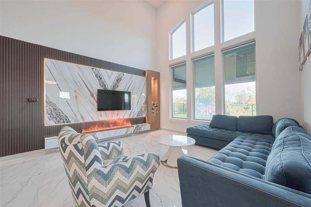 The grand family room with soaring ceilings, massive windows, and a modern electric fireplace with LED lighting and remote ambiance controls creates an inviting gathering space, elevated by intricate wall paneling details.
