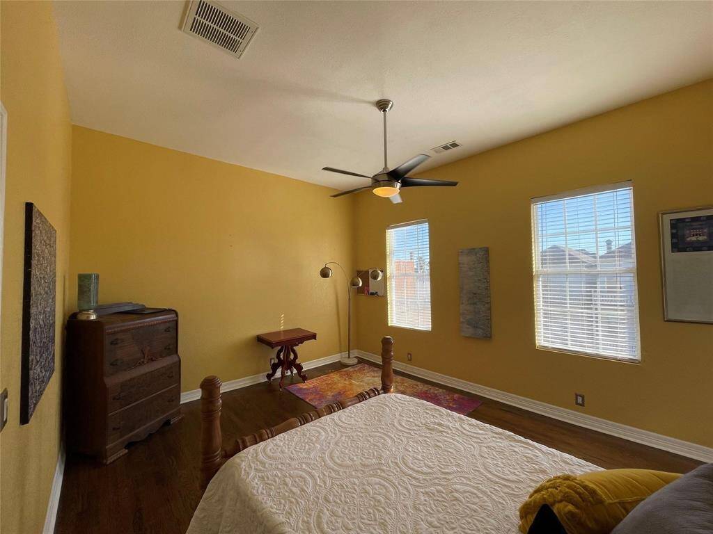 This is a cozy bedroom 2 with warm yellow walls and hardwood flooring. It features a large window with natural light, a ceiling fan, and a colorful area rug. The space includes a bed, a decorative floor lamp, and a small wooden desk.