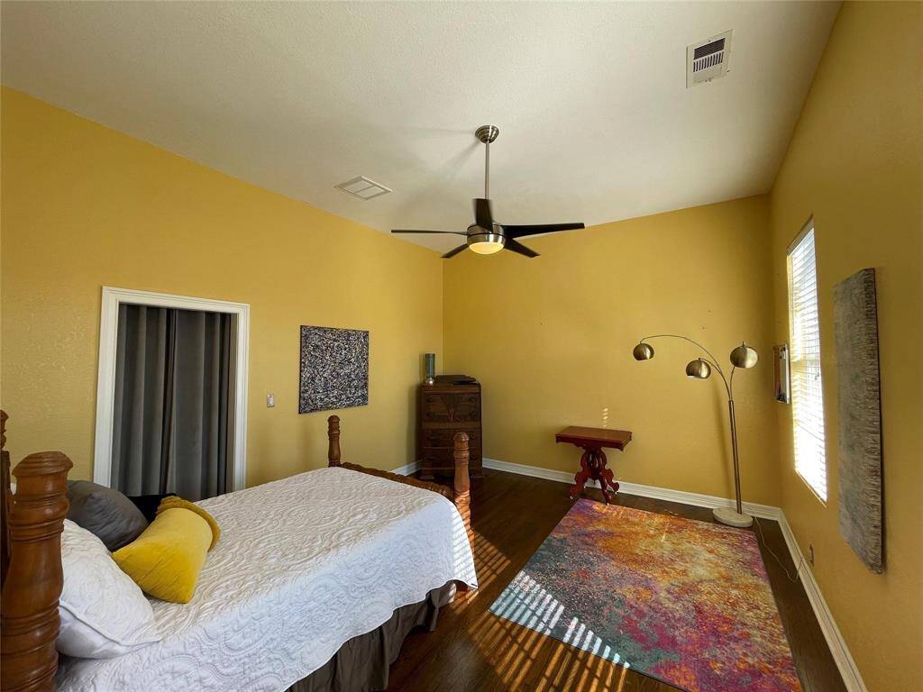 Bright bedroom with warm yellow walls, wood flooring, and ample natural light from two large windows. Features a ceiling fan, a classic dresser, and a queen bed.