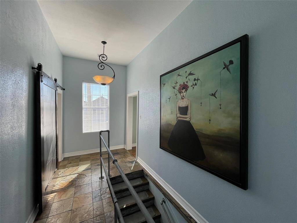 This beautiful hallway entrance features tiled flooring, light blue walls, and high ceilings. It includes a barn-style sliding door, a window allowing natural light, and a decorative hanging light fixture above a staircase with a contemporary metal railing.
