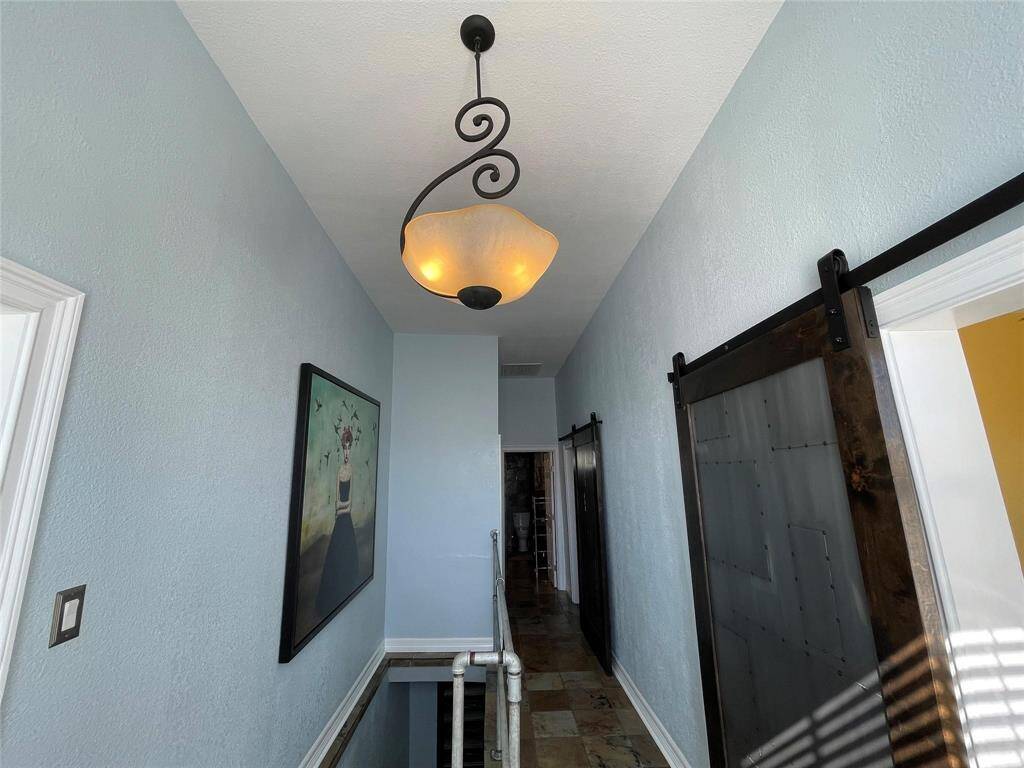 Another view of the hallway entry. This features soft blue walls, a stylish hanging light fixture, and a modern barn door. The space is accented with artwork and has stone tile flooring, creating a contemporary and inviting atmosphere.