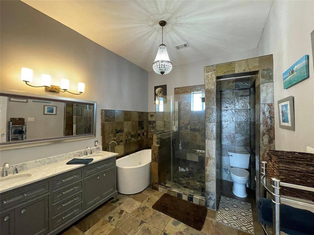 This bathroom features a stylish design with a large double vanity, modern lighting, a freestanding tub, and a glass-enclosed shower. The space is enhanced by stone tile accents and a chandelier, creating a luxurious atmosphere.