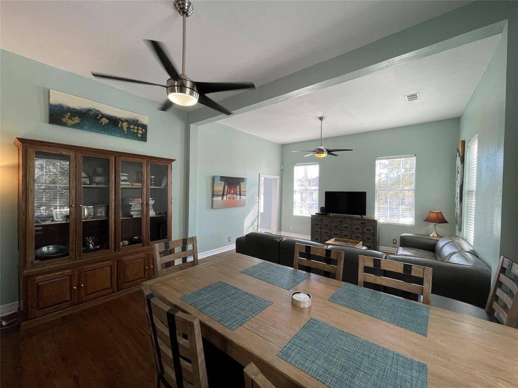 This photo showcases a bright, open-concept living and dining area. It features high ceilings with modern ceiling fans, ample natural light from multiple windows, and a large seating arrangement. The dining area includes a table with matching chairs, while a stylish cabinet provides storage. The walls are painted in a soft green, adding a calming touch to the space.
