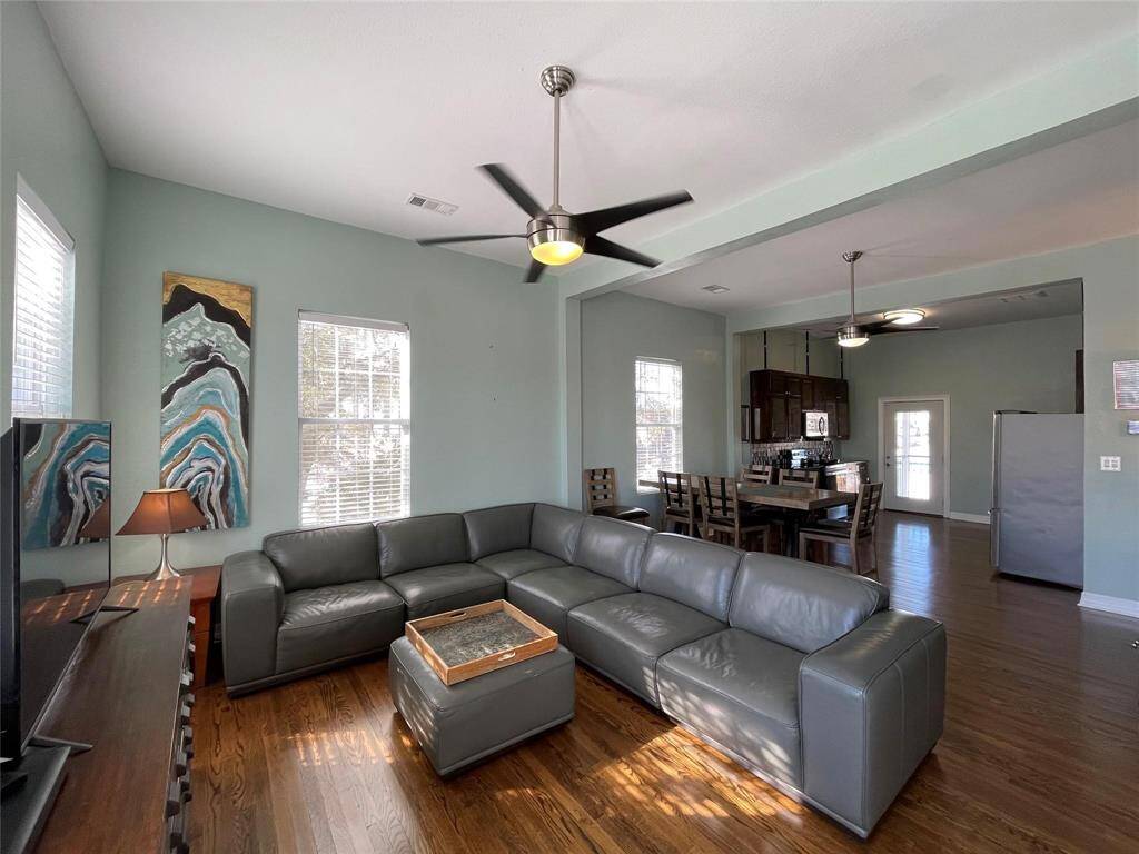 This inviting living space features an open floor plan with abundant natural light, a modern ceiling fan, and hardwood flooring. The room includes a large living area that could comfortably fit a sectional sofa (this sofa not included), contemporary artwork, and a seamless flow into the dining area and kitchen, highlighting both style and functionality.